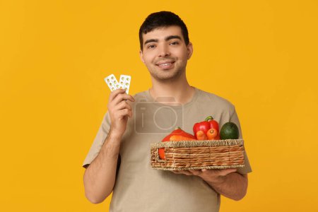 Junger Mann mit Blasen von Vitamin-A-Pillen und gesunder Ernährung auf gelbem Hintergrund