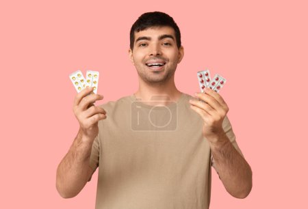 Young man with blisters of vitamin A pills on pink background