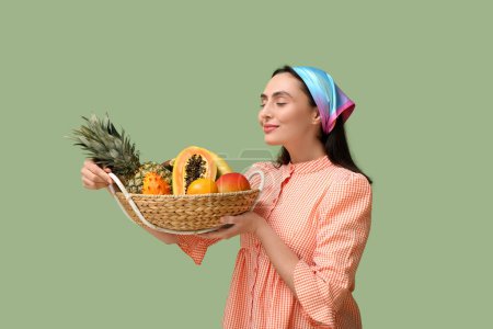 Beautiful young woman holding wicker basket with exotic fruits on green background