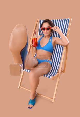 Sexy young woman in swimsuit and with cocktail relaxing on deck chair against beige background
