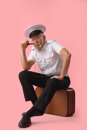 Photo for Thoughtful mature sailor with smoking pipe sitting on bag against pink background - Royalty Free Image