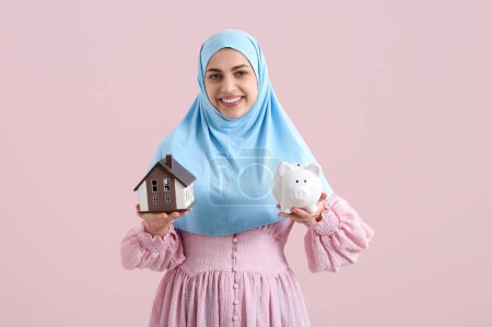 Schöne junge glückliche Muslimin mit Sparschwein und Hausmodell auf rosa Hintergrund. Hypothekenkonzept