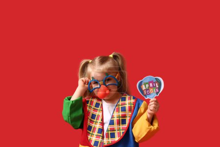 Foto de Niña en traje de payaso con gafas divertidas y tarjeta sobre fondo rojo - Imagen libre de derechos
