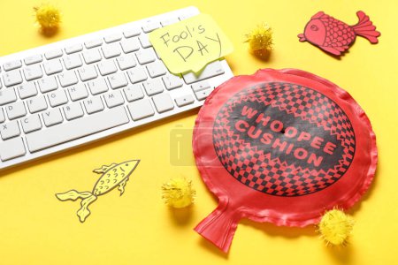 Computer keyboard, sticky note and whoopee cushion on yellow background. April Fools Day prank