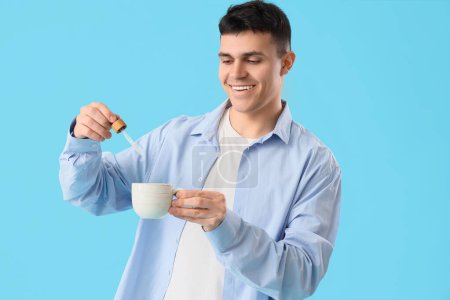 Young man dropping CBD oil into tea cup on blue background