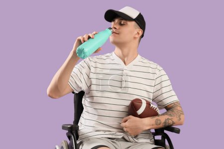 Sporty young man in wheelchair with rugby ball drinking water on lilac background