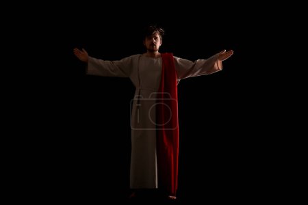Man in Jesus robe and crown of thorns outstretching hands with light on black background