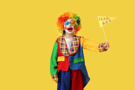 Foto de Niño divertido en traje de payaso con bandera de papel sobre fondo amarillo. Celebración del Día de los Inocentes - Imagen libre de derechos