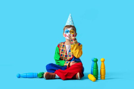 Funny little boy in clown costume with party whistle and juggling clubs on blue background. April Fools' Day celebration