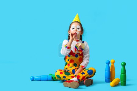 Funny little girl in clown costume with party whistle and juggling clubs on blue background. April Fools' Day celebration