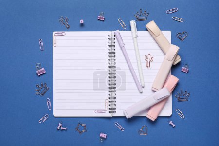 Offenes Notizbuch mit Filzstift, Filzstiften und Büroklammern auf blauem Hintergrund. Ansicht von oben