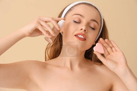 Beautiful young woman with heart-shaped sponge applying makeup foundation on beige background, closeup