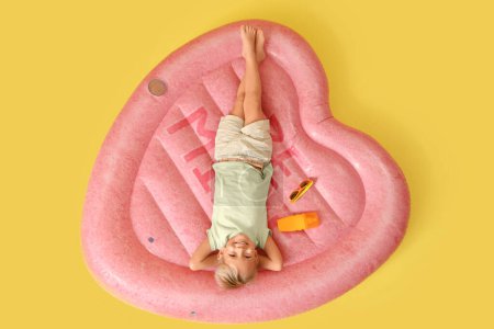 Cute little boy with sunscreen cream lying on swim mattress against yellow background, top view