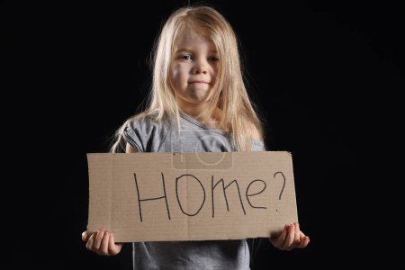 Obdachloses kleines Mädchen mit einem Stück Pappe mit dem Wort HOME auf dunklem Hintergrund
