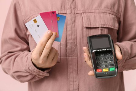 Young man with credit cards and payment terminal on pink background, closeup