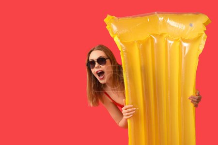 Beautiful surprised young woman in tank swimsuit and with inflatable mattress on red background