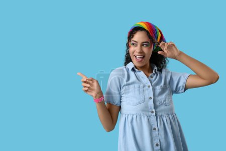 Mujer afroamericana feliz con el pañuelo en la cabeza del arco iris apuntando a algo en el fondo azul. Concepto LGBT