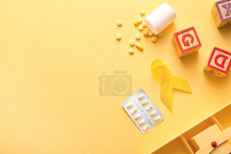 Téléchargez les photos : Ruban doré avec des pilules et des jouets sur fond jaune. Concept de sensibilisation au cancer chez les enfants - en image libre de droit
