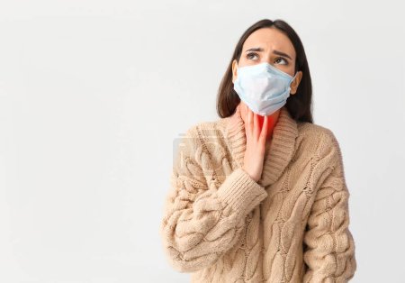 Young woman in medical mask suffering from sore throat on light background
