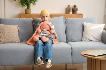 Téléchargez les photos : Triste petite fille après la chimiothérapie avec lapin jouet assis sur le canapé à la maison. Journée internationale du cancer chez les enfants - en image libre de droit