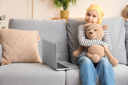 Téléchargez les photos : Petite fille après la chimiothérapie avec ours jouet regarder des dessins animés sur ordinateur portable à la maison. Journée internationale du cancer chez les enfants - en image libre de droit