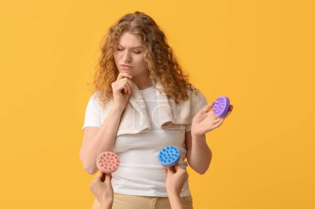 Thoughtful young woman and hands with hair scalp massagers on yellow background