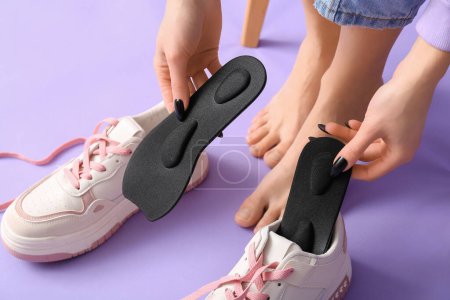 Photo for Young woman putting orthopedic insoles in sneakers on lilac background, closeup - Royalty Free Image