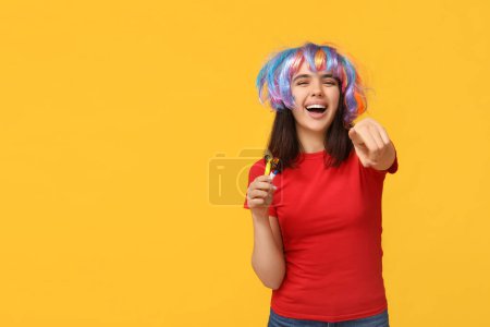 Foto de Hermosa joven disfrazada con silbatos de fiesta apuntando al espectador sobre un fondo amarillo. Celebración del día del tonto de abril - Imagen libre de derechos
