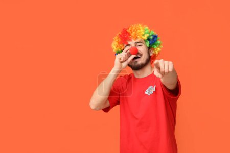 Handsome young man in funny clown disguise pointing at viewer on orange background. April fool's day celebration