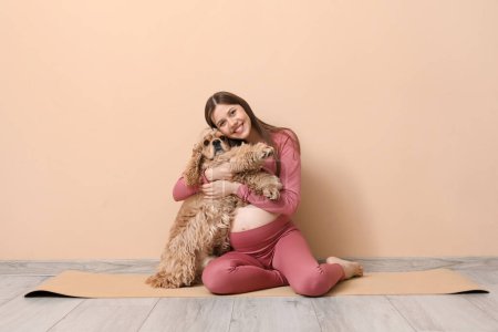 Photo for Beautiful young sporty pregnant woman with cocker spaniel hugging near beige wall - Royalty Free Image