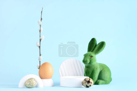 Podios decorativos con huevos de Pascua, conejito de juguete y rama de sauce sobre fondo azul