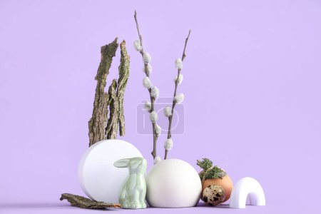 Podios decorativos con huevos de Pascua, conejito de juguete y ramas de sauce sobre fondo lila
