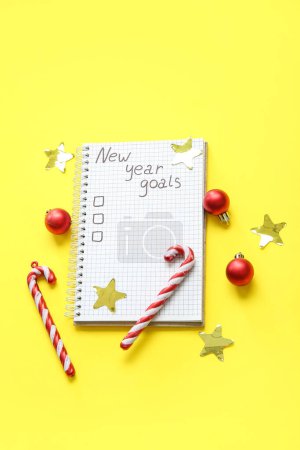 Foto de Cuaderno con lista de tareas vacías, bastones de caramelo y bolas de Navidad sobre fondo amarillo. Objetivos de año nuevo - Imagen libre de derechos