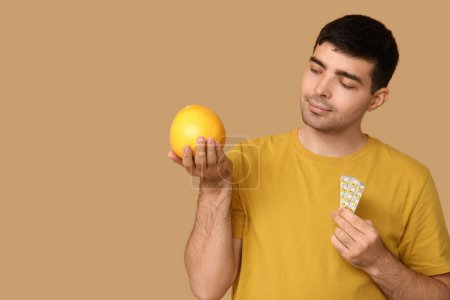 Young man with blisters of vitamin A pills and grapefruit on beige background