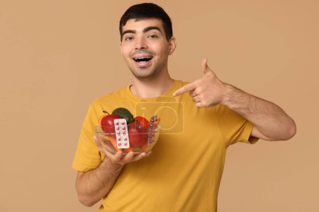 Foto de Hombre joven apuntando a las píldoras de vitamina A y alimentos saludables en fondo beige - Imagen libre de derechos