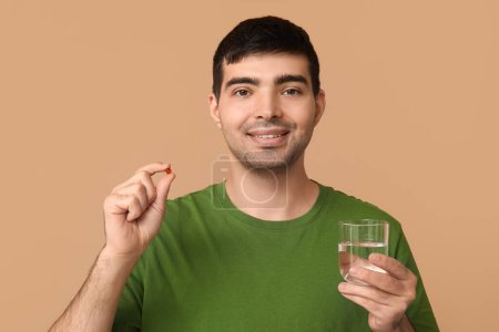 Young man with vitamin A pill and glass of water on beige background, closeup