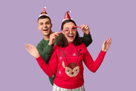 Foto de Pareja joven divertida en pijamas de Navidad y con bastones de caramelo sobre fondo lila - Imagen libre de derechos