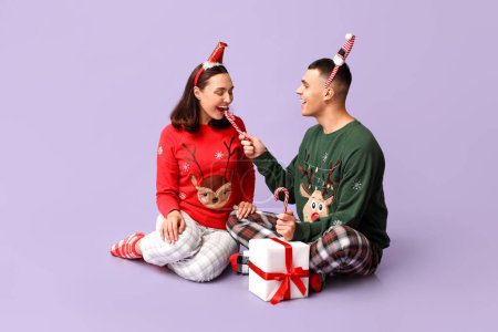 Foto de Feliz pareja joven en pijama de Navidad, con bastones de regalo y dulces sobre fondo lila - Imagen libre de derechos