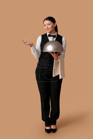 Téléchargez les photos : Belle jeune serveuse avec plateau et cloche sur fond marron - en image libre de droit