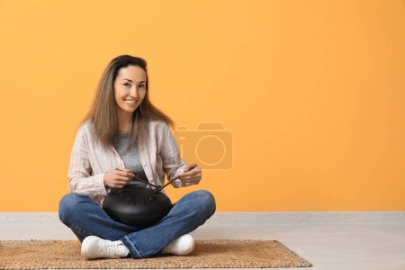 Schöne reife Frau mit Stöcken spielt Glukophonie und sitzt auf Teppich in der Nähe der gelben Wand