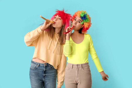 Schöne junge Frauen in lustigen Clownskostümen mit Party-Trillerpfeifen auf blauem Hintergrund. Feier zum Aprilscherz
