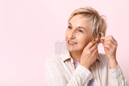 Mujer madura poniendo audífono en su oreja sobre fondo rosa