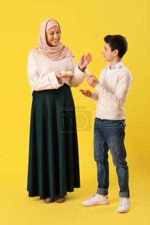 Happy Muslim mother and her son with traditional sweets on yellow background. Eid al-Fitr celebration