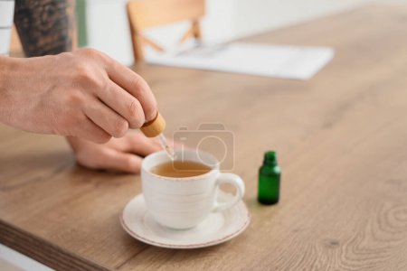 Young man dropping CBD oil into tea cup in kitchen, closeup