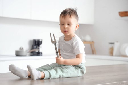 Little boy with sharp meat fork sitting on table in kitchen. Child at risk