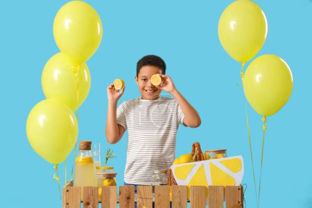 Little African-American boy with lemons at lemonade stand on blue background