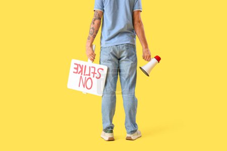 Photo for Protesting young man holding placard with text ON STRIKE and megaphone against yellow background, back view - Royalty Free Image