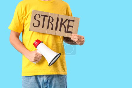 Photo for Protesting young man holding placard with word STRIKE and megaphone on blue background, closeup - Royalty Free Image