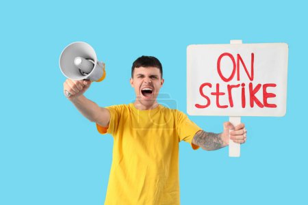 Photo for Protesting young man holding placard with text ON STRIKE and megaphone against blue background - Royalty Free Image