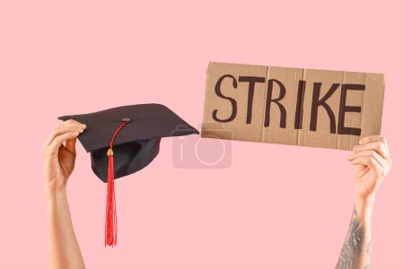 Protesting male graduate holding placard with word STRIKE and mortar board on pink background